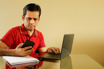 Adult man sitting at desk in living room of his house, with yellow background wall, operating his notebook and looking at his smartphone