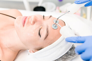 Naklejka premium The young female client of cosmetic salon having microcurrent procedure on her face with special devices, close-up. Beautician using electrical impulses for facial procedures