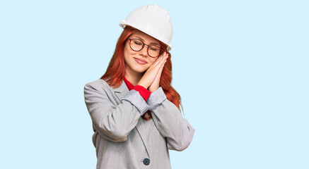Young redhead woman wearing architect hardhat sleeping tired dreaming and posing with hands together while smiling with closed eyes.