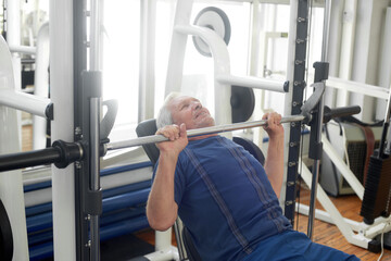 Fototapeta na wymiar Elderly male is having intense workout in gym. Senior caucasian man lifting weights at fitness club. Strength workout for seniors.