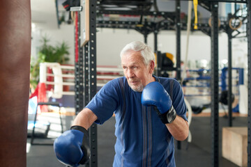 Senior man training with a punching bag. Older concentrated man boxing punching bag at gym. People,...