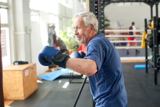 Senior man beats punching bag in gym. Male pensioner prepared for a fight with blue boxing gloves at boxing hall. Elderly man in gym.