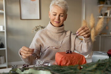 Attractive female pensioner in turtleneck sweater holding decorative tape, wrapping Christmas presents, sitting at table with natural fir branches and ornaments, having joyful facial expression