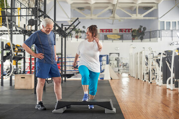 Obraz na płótnie Canvas Joyful elderly woman exercising at gym. Senior man looking at woman during training at gym. Stay active and healthy in any age.