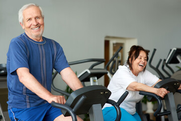 Obraz premium Elderly man working out on exercise bike in gym. Retired caucasian man looking at camera while exercising at fitness club. Training for long living.