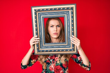 Fototapeta premium Young caucasian woman with blond hair holding empty frame skeptic and nervous, frowning upset because of problem. negative person.