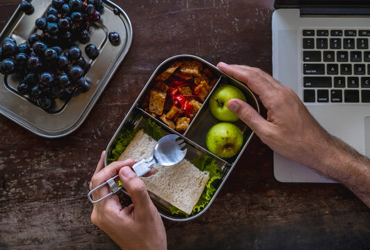 Close up of man hands at desk with a laptop at home or in office with healthy vegan meal in reusable stainless steel lunch box. Healthy lifestyle, zero waste and sustainable plastic free lifestyle