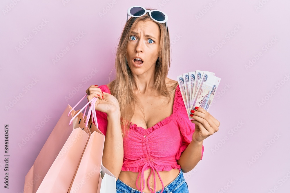 Sticker Beautiful caucasian woman holding shopping bags and 500 russian ruble in shock face, looking skeptical and sarcastic, surprised with open mouth