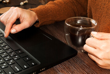 Fototapeta na wymiar Home Office. A woman sits at a laptop and holds a cup of coffee. Glass cup in the shape of a heart and double glass in a female hand. Work at home.