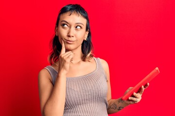 Naklejka premium Young woman holding touchpad tablet serious face thinking about question with hand on chin, thoughtful about confusing idea