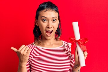 Young woman holding graduate degree diploma pointing thumb up to the side smiling happy with open mouth