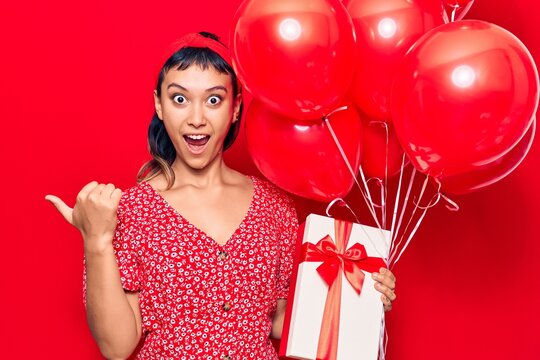 Young woman holding balloons with gift pointing thumb up to the side smiling happy with open mouth
