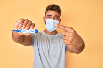 Young hispanic man wearing medical mask holding thermometer smiling happy pointing with hand and finger