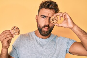 Young hispanic man holding cookie making fish face with mouth and squinting eyes, crazy and comical.