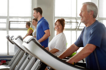 Senior woman running on treadmill smiling at camera. People working out at fitness center.