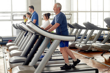 People working out at gym. Senior man walking on treadmill at modern fitness club. Cardio workout...