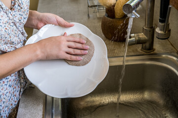 Woman hands cleaning plate with natural compostable hemp dish washing sponge by sink with running tap water. Eco-friendly kitchen and home products. Zero waste sustainable plastic free lifestyle