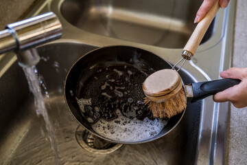 Woman hands cleaning frying pan with natural compostable dish washing brush by the sink with running tap water. Eco-friendly kitchen and home products. Zero waste sustainable plastic free lifestyle