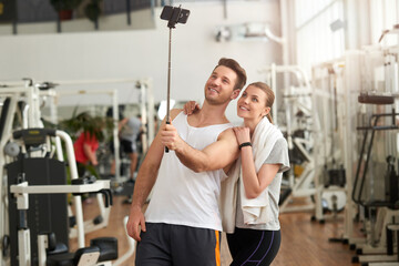 Fototapeta na wymiar Athletic couple using selfie stick at gym. Young beautiful couple taking selfie with monopod at modern fitness club. People, sport, leisure, lifestyle.