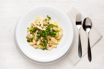 Pasta with broccoli and chicken on a white plate on a white wooden table with a spoon and fork