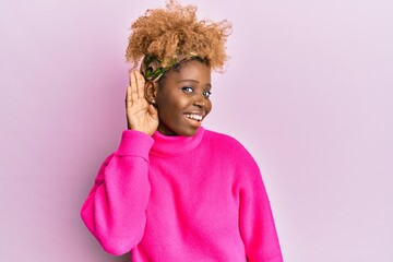 Young african woman with afro hair wearing casual winter sweater smiling with hand over ear listening and hearing to rumor or gossip. deafness concept.