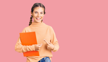 Beautiful caucasian woman with blonde hair holding book smiling happy pointing with hand and finger