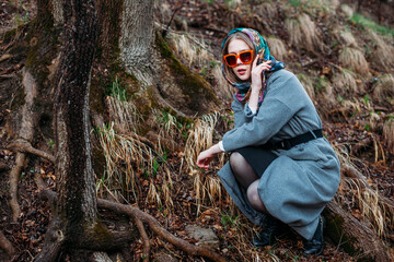 fashion shooting. young model wearing bright silk headscarf and orange-rimmed sunglasses sits under tree in an autumn gray forest