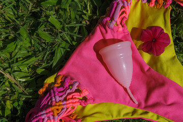 Silicone menstrual cup with pink bikini swimsuit on grass. Reusable eco-friendly feminine hygiene products during summer vacation. Zero waste sustainable plastic free lifestyle
