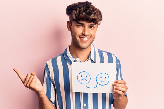 Young hispanic man holding sad to happy emotion paper smiling happy pointing with hand and finger to the side