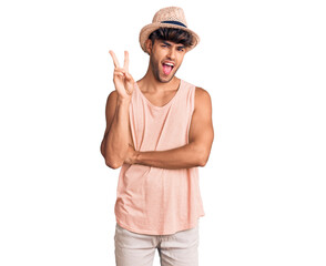Young hispanic man wearing summer hat smiling with happy face winking at the camera doing victory sign. number two.