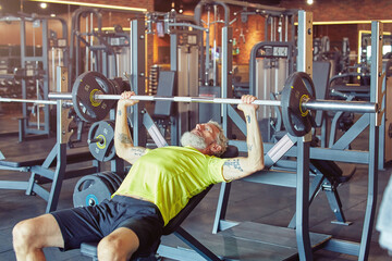 Working hard. Strong mature man in sportswear lifting weighted bar or barbell at gym