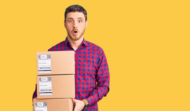 Handsome young man with bear holding delivery packages scared and amazed with open mouth for surprise, disbelief face