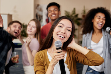 Bring joy. Young pretty asian woman holding microphone and singing while having fun with friends at...