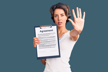 Young beautiful woman holding clipboard with agreement document with open hand doing stop sign with serious and confident expression, defense gesture