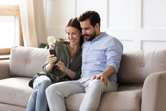 Smiling young man and woman sit relax on couch in living room look at cellphone screen make self-portrait picture together. Happy Caucasian couple rest on sofa at home use smartphone gadget.