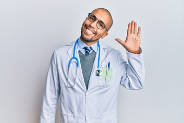 Hispanic adult man wearing doctor uniform and stethoscope waiving saying hello happy and smiling, friendly welcome gesture