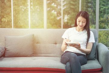 Young beautiful Asian woman relaxing in living room at home, reading and writing on book in the morning with happy feeling,workation concept.