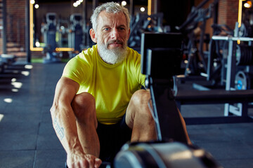 Working out in fitness club. Strong mature athletic man in sportswear exercising on rowing machine at gym