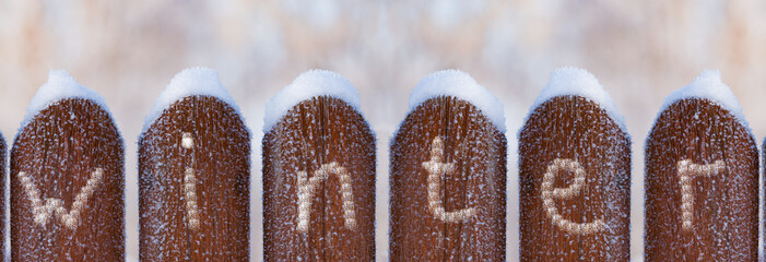Fence made of dark brown boards in frost and snow with the inscription winter. Soft blurry background, a copy of the space. Beautiful winter background