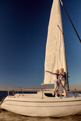 Fototapeta premium Sailing around world. Full length of happy senior couple standing on the side of sail boat or yacht deck floating in sea and enjoying amazing sunset, vertical shot