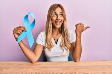 Beautiful blonde young woman holding blue ribbon pointing thumb up to the side smiling happy with...