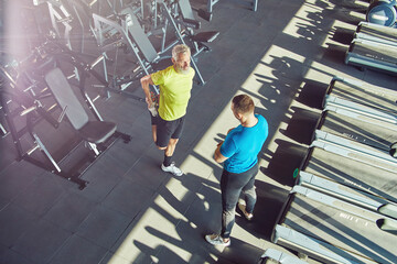 Middle aged man in sportswear warming up, stretching his legs and talking with fitness instructor at gym or sport club, full length shot