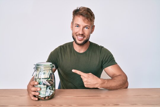 Young caucasian man holding jar with savings sitting on the table smiling happy pointing with hand and finger