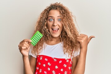 Beautiful caucasian teenager girl holding birth control pills pointing thumb up to the side smiling happy with open mouth