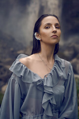 portrait of a woman in a gray dress stands against a high waterfall in summer