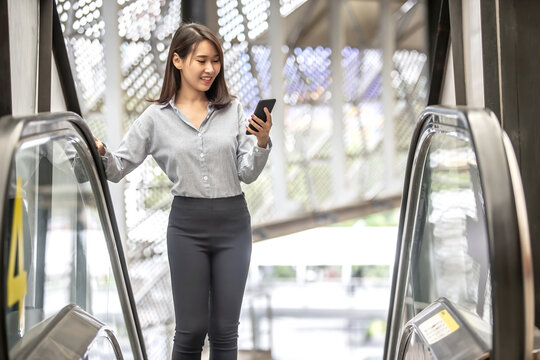 Young Asian Business Woman Using Her Smartphone On The Escalator In A Shopping Mall