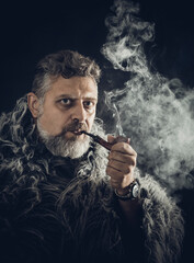 Solid bearded man in a fur mantle smoking pipe