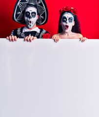 Young couple wearing mexican day of the dead costume holding blank empty banner scared and amazed with open mouth for surprise, disbelief face