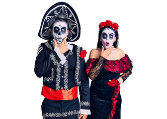 Young couple wearing mexican day of the dead costume over background looking fascinated with disbelief, surprise and amazed expression with hands on chin
