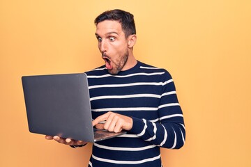 Young handsome man working using laptop standing over isolated yellow background scared and amazed...